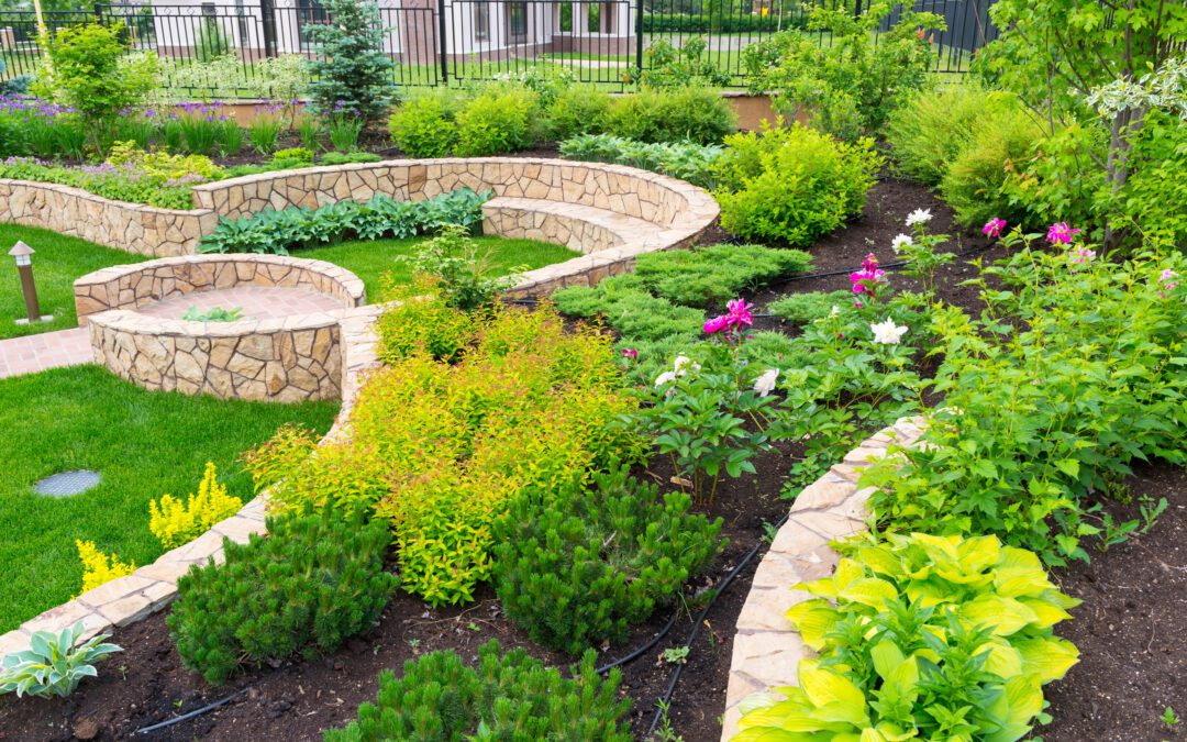 A well-maintained residential garden featuring tiered landscaping with stone walls, lush green lawn areas, and various plants including shrubs, flowering plants, and small trees.
